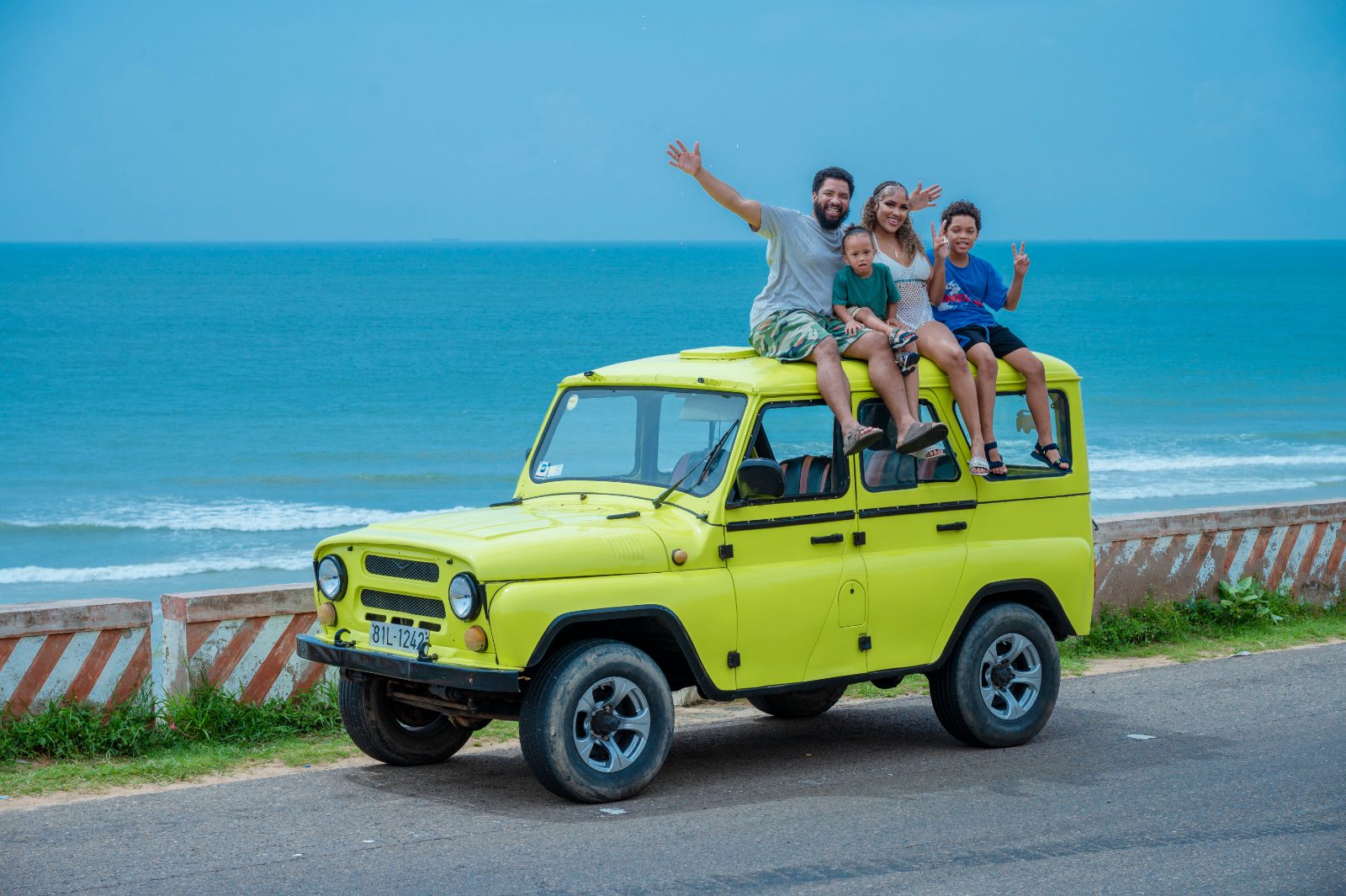 Marcellous, Keana, and their two boys waving from the roof of a yellow jeep by the sea
